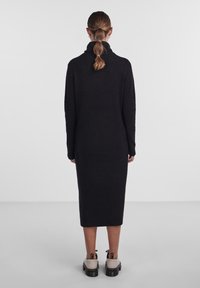 Vestito di lana nera con colletto alto, maniche lunghe e silhouette dritta, che arriva a metà polpaccio. Il modello indossa scarpe beige chiaro.