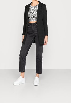 Person iført en sort blazer, hvid og sort zebramønstret crop top, sorte højtaljede jeans og hvide sneakers, stående mod en ensfarvet baggrund.
