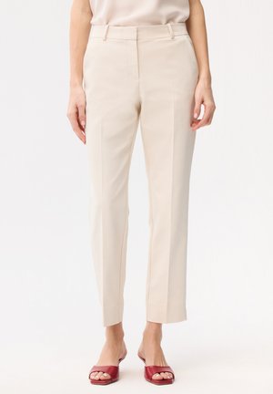 Vrouw draagt beige getailleerde broek en rode open-teen slip-on hakken, staand tegen een effen witte achtergrond.