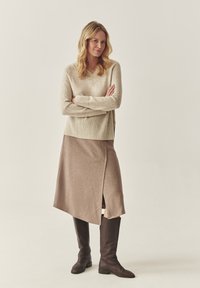 Beige kötött pulóver V-nyakkal és bordás mandzsettákkal, egy bézs aszimmetrikus szoknyával és barna térdharisnyás csizmával párosítva. Egyszerű és modern dizájn.