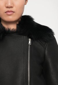 Artimas kadras asmens, vilkinčio juodą odinę striukę su sidabriniu užtrauktuku ir pūkuotu juodu kailiniu apykakle, esantis prieš vienodą šviesią foną.