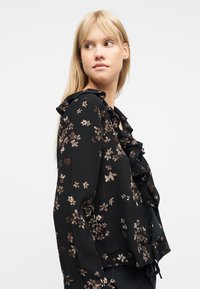 Νεαρή γυναίκα με ξανθά μαλλιά που φοράει μαύρο μπλουζάκι με λουλουδάτο σχέδιο και βολάν, κοιτάζοντας πλάγια σε λευκό φόντο.