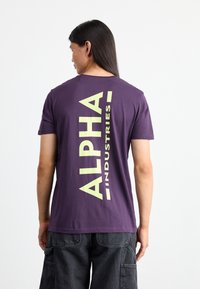 Lila pamut póló, hátul nagy, függőleges sárga felirattal, amely így olvasható: "ALPHA INDUSTRIES". Rövid ujjak és szabványos fit.