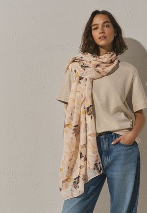 Jeune femme aux cheveux mi-longs porte un haut beige à manches courtes, une écharpe florale ample et un jean bleu avec une main dans la poche.