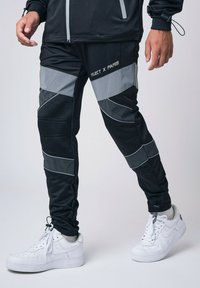 Pantalones deportivos negros con acentos de malla gris y paneles reflectantes. Presentan un corte ajustado y puños elásticos. Combinados con zapatillas blancas.