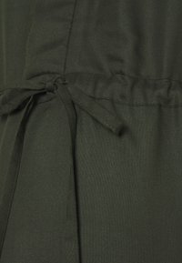 Mörkgrön tyg med en slät struktur och en midja som är rynkad, med ett knytbart bälte för justerbar passform och en enkel, nedtonad design.