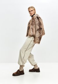Kvinde iført en brun faux fur jakke, beige stribede løse bukser, brune sko og står imod en ensfarvet lys baggrund.