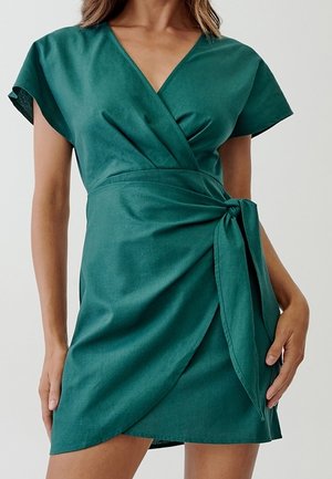 Kobieta w zielonej sukience kopertowej z krótkim rękawem, dekoltem w serek i wiązanym paskiem w talii.