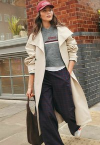 Beige trenchcoat over een grijze trui met rode opdruk, gecombineerd met brede marineblauwe geruite broek en witte sneakers. Bruine tote bag in de hand.