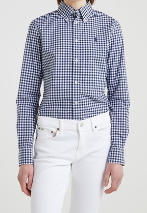 Osoba ubrana w dopasowaną, niebiesko-białą koszulę w kratę z długimi rękawami, połączoną z białymi, wysokim stanem spodniami.