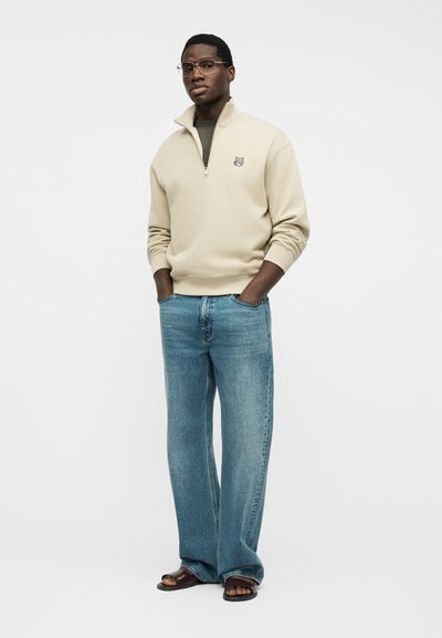 Άνδρας που φοράει μπεζ μπλούζα με φερμουάρ μέχρι το στήθος και λογότυπο λύκου, ανοιχτόχρωμα μπλε τζιν, καφέ σανδάλια και γυαλιά, στέκεται με τα χέρια στις τσέπες.