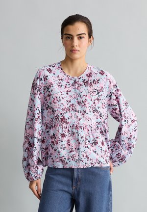 Junge Frau mit dunklen Haaren trägt eine langärmelige rosa-lila Bluse mit Blumenmuster und blaue Jeans vor schlichtem grauem Hintergrund.