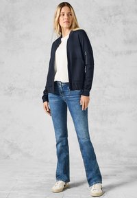 Giacca compressibile blu navy con maniche lunghe, due tasche frontali e una tasca con zip sul braccio, abbinata a jeans blu a zampa d'elefante e sneakers bianche.