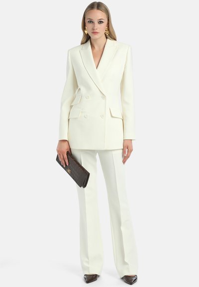 Witte dubbelknop blazer met twee voorzakken, gecombineerd met op maat gemaakte witte flared broeken en een donkerbruine gestructureerde clutch.