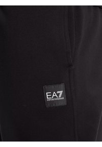 Pantalons de survêtement noirs en tissu doux, dotés d'un patch logo EA7 Emporio Armani brodé sur le côté. Design simple avec poches.