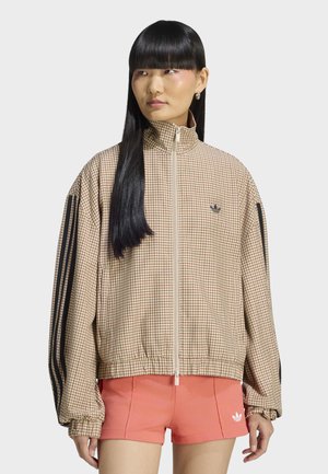 Kvinde med lige sort hår iført beige ternet lynlåstrøje med sorte striber på ærmerne og koralfarvede shorts med hvidt logo.