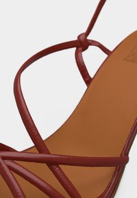 Κοντινό πλάνο ενός καφέ σανδαλιού με λεπτές, γυαλιστερές κόκκινες λωρίδες που διασταυρώνονται πάνω σε ταμπά πάτο.