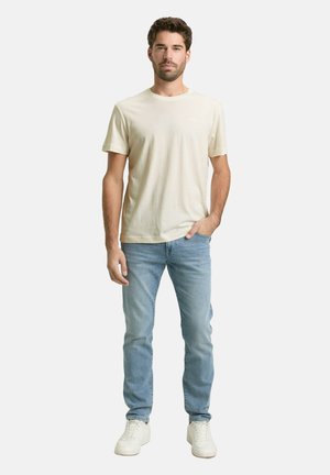 Man met kort haar en baard staand, gekleed in een crème korte mouw t-shirt, lichtblauwe spijkerbroek, witte sneakers, één hand in de broekzak.