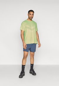 Ανοιχτό πράσινο αθλητικό μπλουζάκι κοντό μανίκι με υφή, συνδυασμένο με ναυτικό μπλε βερμούδα και μαύρα αθλητικά παπούτσια και κάλτσες.