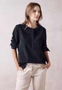 Camicia nera con colletto tondo e maniche arrotolate, abbinata a pantaloni beige chiaro con coulisse; texture liscia, vestibilità casual.