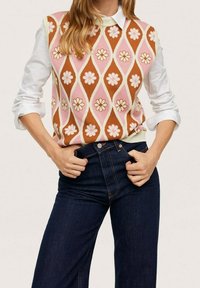 Strickweste mit einem rosa, braunen und weißen Blumendesign, über einem weißen Hemd mit Kragen getragen; kombiniert mit dunkelblauen Jeans.