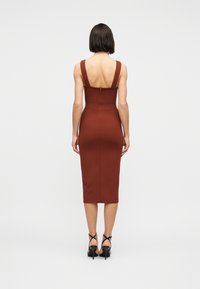 Γυναίκα με κοντά σκούρα μαλλιά, φοράει αμάνικο, εφαρμοστό midi φόρεμα σε σκουριασέ απόχρωση και μαύρα πέδιλα με τακούνι, ενώ στέκεται γυρισμένη ανάποδα.