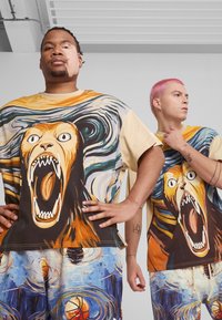 To personer bærer oversized grafiske t-shirts med et brølende løve design i varme nuancer, kombineret med bukser med abstrakte mønstre i blå og orange.