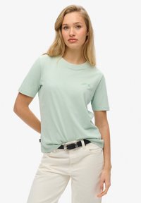 Camiseta de algodón verde claro con mangas cortas, cuello redondo y un sutil logotipo bordado. Llevada con pantalones blancos y un cinturón negro.