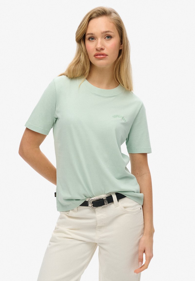 Camiseta de algodón verde claro con mangas cortas, cuello redondo y un sutil logotipo bordado. Llevada con pantalones blancos y un cinturón negro.