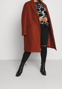 Abrigo marrón de mezcla de lana, de doble botonadura, con grandes solapas, llevado sobre un suéter gráfico negro y jeans ajustados rasgados negros, combinado con botas de tacón negras.