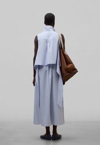 Käppade juustega inimene, kes kannab helesinist varrukateta kleiti ja tumedaid libisevaid kingi, õlal suur pruun käekott.