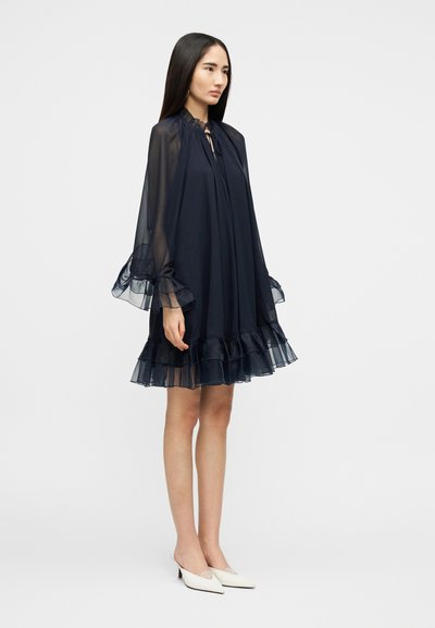 Γυναίκα που στέκεται, φορώντας ένα διάφανο, μακρυμάνικο φόρεμα σε ναυτικό μπλε με βολάν στα μανίκια και στο τελείωμα, συνδυασμένο με άσπρα μυτερά γοβάκια με χαμηλό τακούνι.