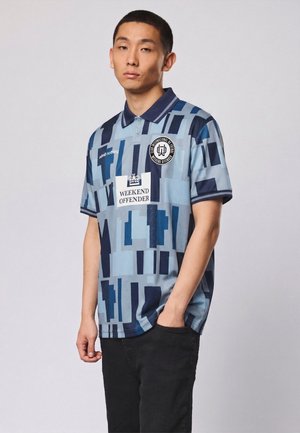 Nuori mies, jolla on yllään sininen kuvioitu polo-paita, jossa on "Weekend Offender" -logo, seisoo mustien housujen taskuun yhdellä kädellä tyhjää taustaa vasten.