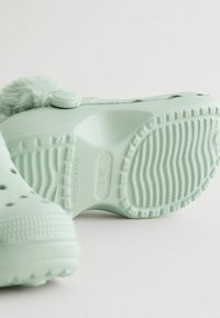 Sabots vert clair avec une surface texturée, un accent moelleux et une semelle à motifs. Dotés d'un bouton circulaire pour l'ajustement.