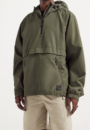 Homme portant une veste anorak à capuche vert olive avec poches avant et un short beige, debout devant un fond clair uni.