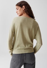 Femme aux cheveux châtain ondulés portant un pull texturé vert clair et un jean gris foncé, se retournant sur son épaule devant un fond uni.