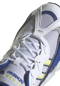 Baskets blanches de sport avec tige en mesh, accents bleus et argentés, six œillets et rayures jaunes. Présente une surface texturée et un système de laçage.