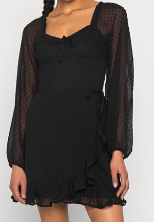 Vestido largo negro transparente de manga larga con textura de puntos, escote corazón, detalle de lazo en el frente, falda cruzada y bajo con volantes, llevado por una persona.