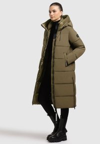 Lang olijfgroene pufferjas met capuchon, rits aan de voorkant en geribbelde manchetten. Kenmerkt zich door een soepele textuur en gewatteerd ontwerp. Gekleed met zwarte legging en laarzen.