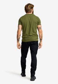 Olivgrön t-shirt med kort ärm och en figurnära design, bärs med svarta cargo-byxor och mörka skor. Tatuerade armar syns från baksidan.