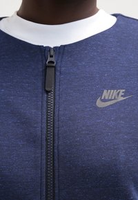 Marinblå zip-up-jacka i texturerat tyg, med vit krage och en liten grå Nike-logga på vänster sida. Svart dragkedja.