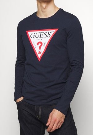 Osoba w granatowej koszuli z długim rękawem z dużym czerwonym trójkątnym logo z napisem „GUESS U.S.A. Washed Jeans” oraz ciemnoniebieskich dżinsach.