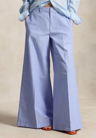 Άτομο που φοράει ανοιχτό μπλε παντελόνι φαρδύ πόδι και αντίστοιχο πουκάμισο, με τα χέρια στις τσέπες, φορώντας ορατά πορτοκαλί σανδάλια σε μπεζ φόντο.