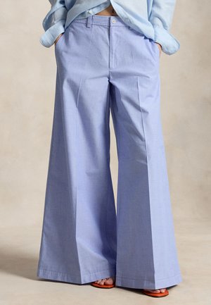 Person som har på seg lyseblå bukser med vide ben og matchende skjorte, med hendene i lommene, og synlige oransje sandaler på beige bakgrunn.