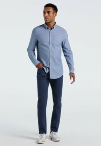 Helles blaues Langarmhemd mit Knopfleiste und Tasche, kombiniert mit marineblauen Hosen und weißen Sneakern mit roten Akzenten. Glatte Stofftextur.