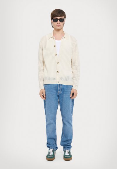 Cream-colored ribbed cardigan with gold buttons, worn over a white tank top. Paired with blue jeans and green sneakers. Black sunglasses.