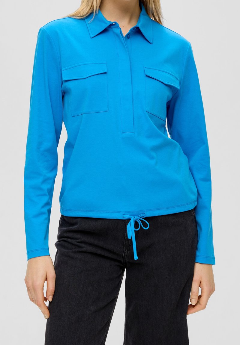Haut à manches longues bleu vif avec col, deux poches poitrine et ourlet à cordon de serrage pour un ajustement personnalisé. Texture de tissu douce.