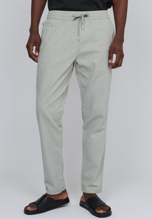 Homme portant un pantalon gris clair avec cordon et des sandales noires ouvertes à l’avant, debout devant un fond uni.