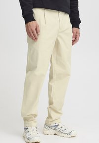 Pantaloni chino bej cu față plisată, croială lejeră și buzunare laterale, asortați cu adidași albi și gri. Material fin și design drept al pantalonilor.