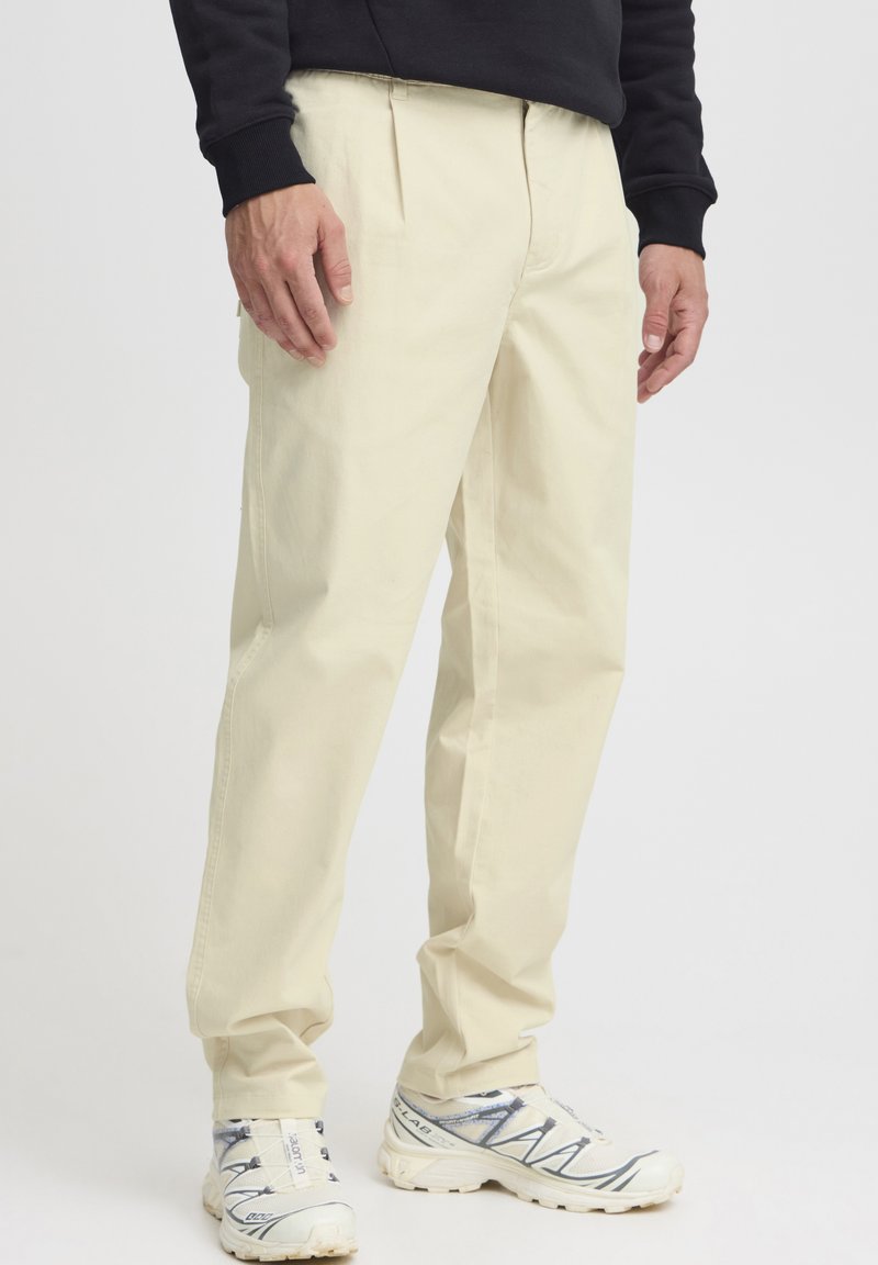 Pantaloni chino bej cu față plisată, croială lejeră și buzunare laterale, asortați cu adidași albi și gri. Material fin și design drept al pantalonilor.